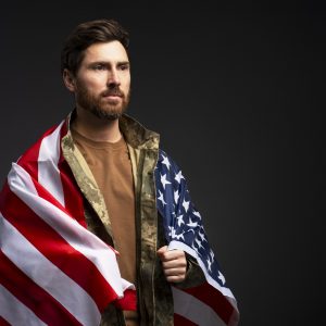 Handsome bearded man, serious confident soldier wearing military camouflage uniform holding American flag isolated on black background, copy space. Independence Day concept