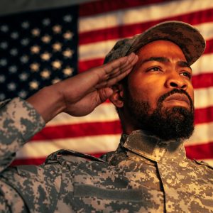 A young adult African American military officer is in the army base, saluting in front of an American flag.