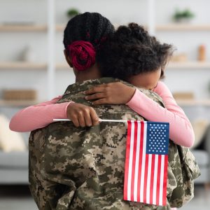 Emotional teen black girl with flag of the US hugging her mom wearing camouflage uniform, daughter resting on her mother shoulder, woman coming back home from military service, back view