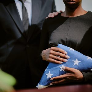 Black woman holding folded American flag standing beside Caucasian man in suit during funeral ceremony outdoors, man resting hand on woman's shoulder, both facing forward